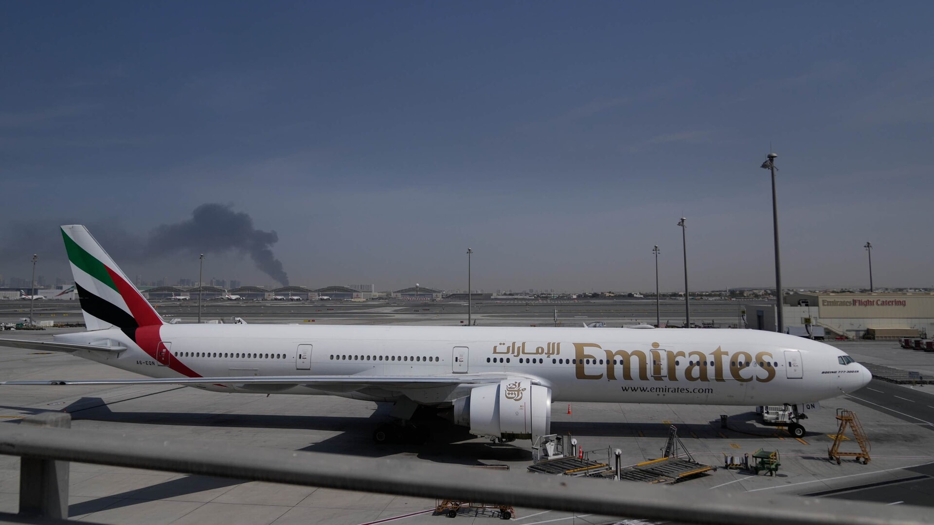 A plume of smoke caused by an Iranian strike is seen in the background as an Emirates plane is parked at Dubai International Airport after its closure